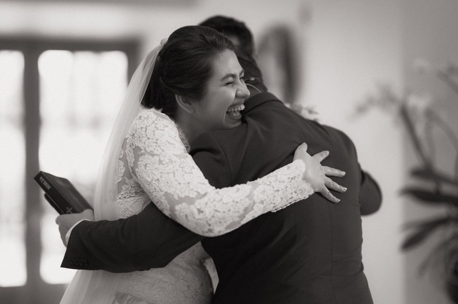 Black and white photo of a bride in lace dress joyfully embracing a guest at her wedding ceremony.