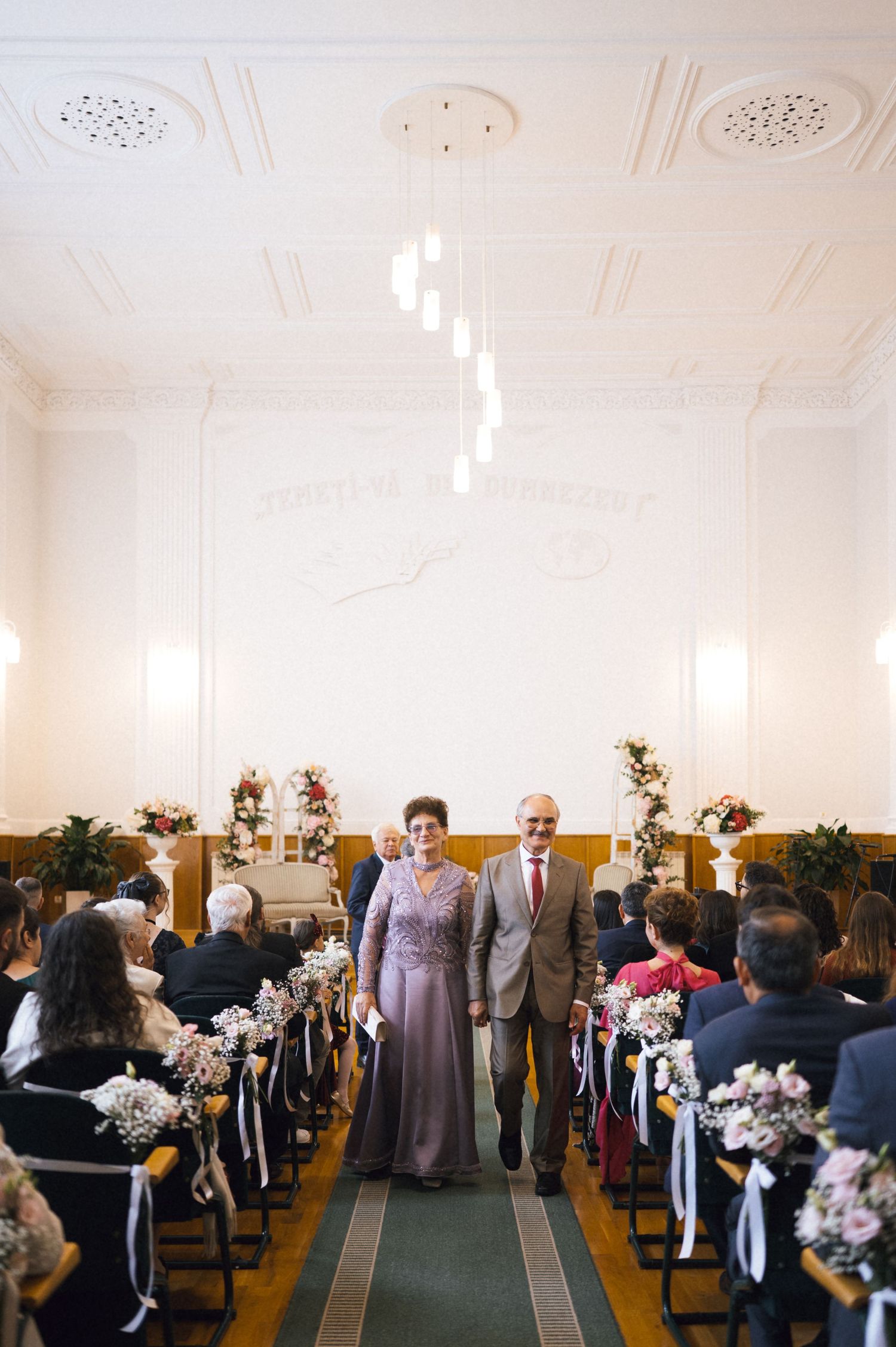 Elegant wedding ceremony in a classic hall with couple walking down the aisle, floral decorations, and seated guests.