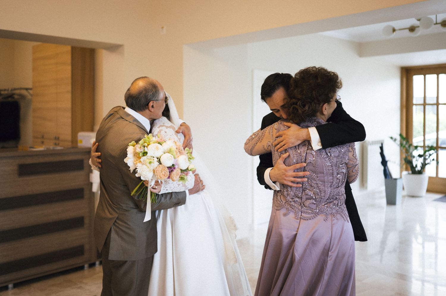 Wedding couple embracing family members indoors, bride holding pink bouquet, emotional family reunion moment.