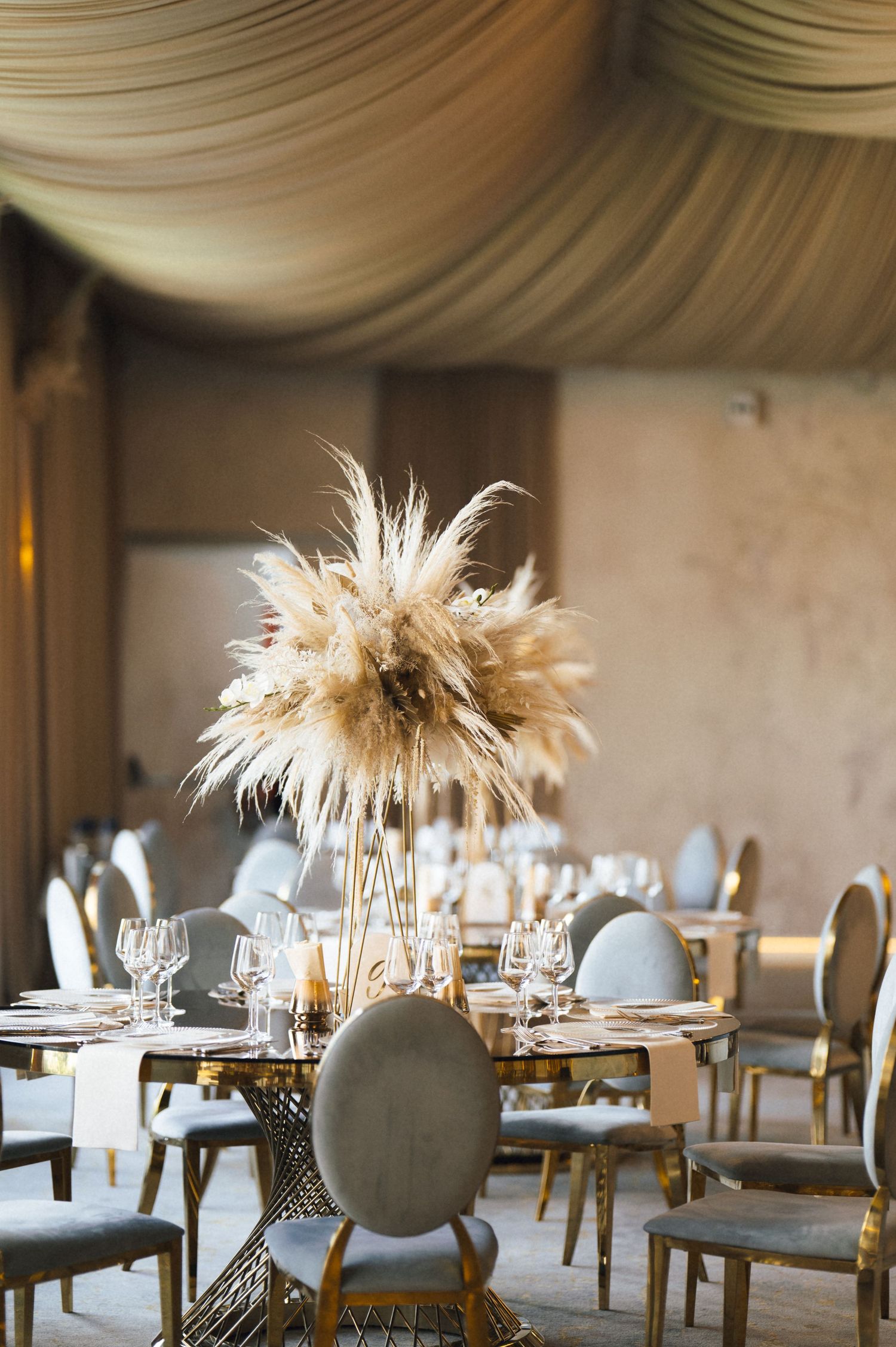 Elegant wedding reception with pampas grass centerpiece on gold table, gray velvet chairs, and draped ceiling decor.