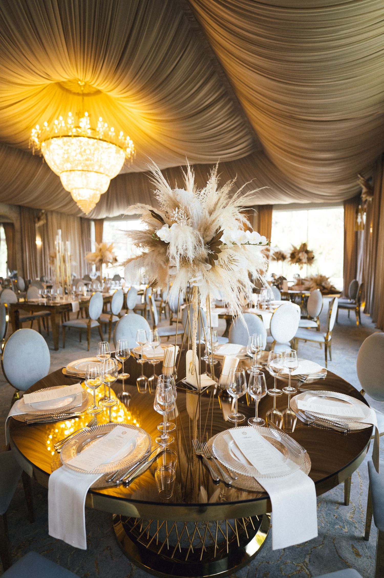 Elegant wedding reception with gold mirror tables, pampas grass centerpieces, crystal chandelier, and draped ivory ceiling decor.