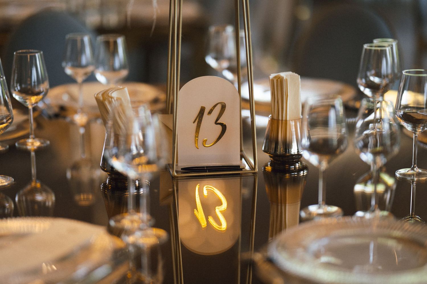 Elegant wedding table setting with gold number 13 centerpiece, crystal glassware, and glowing candle accent.