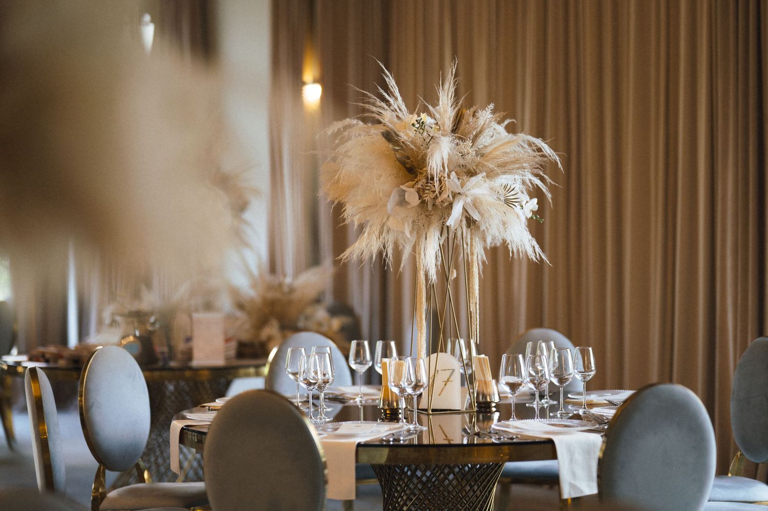 Elegant wedding reception table with tall pampas grass centerpieces, crystal glasses, and gray velvet chairs in a warmly lit ballroom.