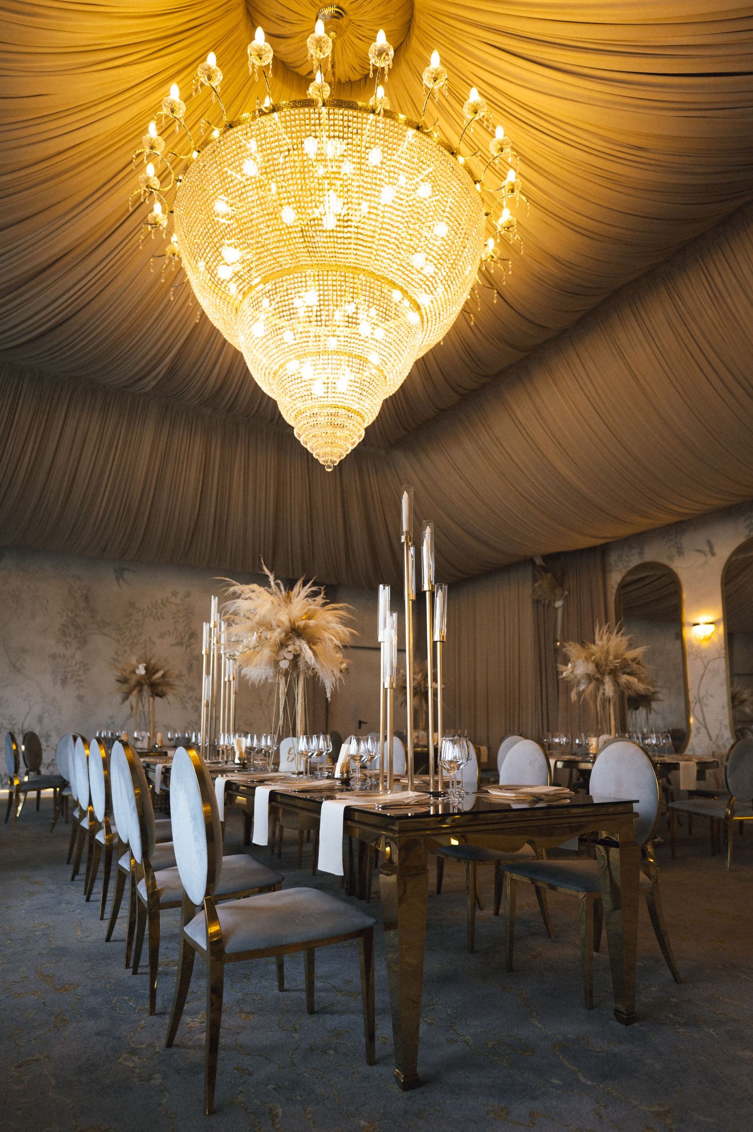 Elegant event venue with a grand crystal chandelier, draped gold ceiling, long dining table, and tall feather centerpieces.