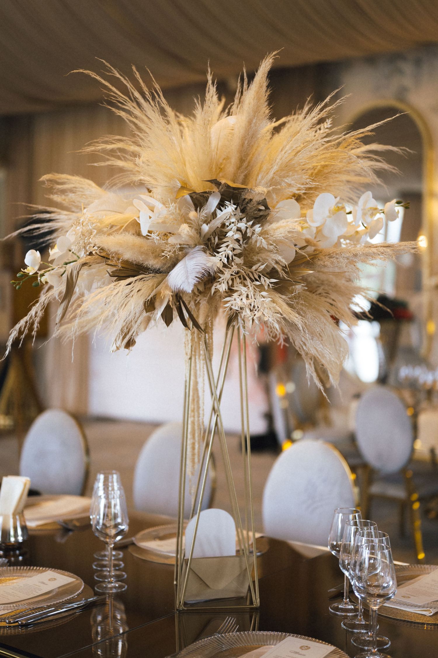 Tall gold geometric vase centerpiece with lush pampas grass and white orchids on a mirrored table at a luxury wedding reception.
