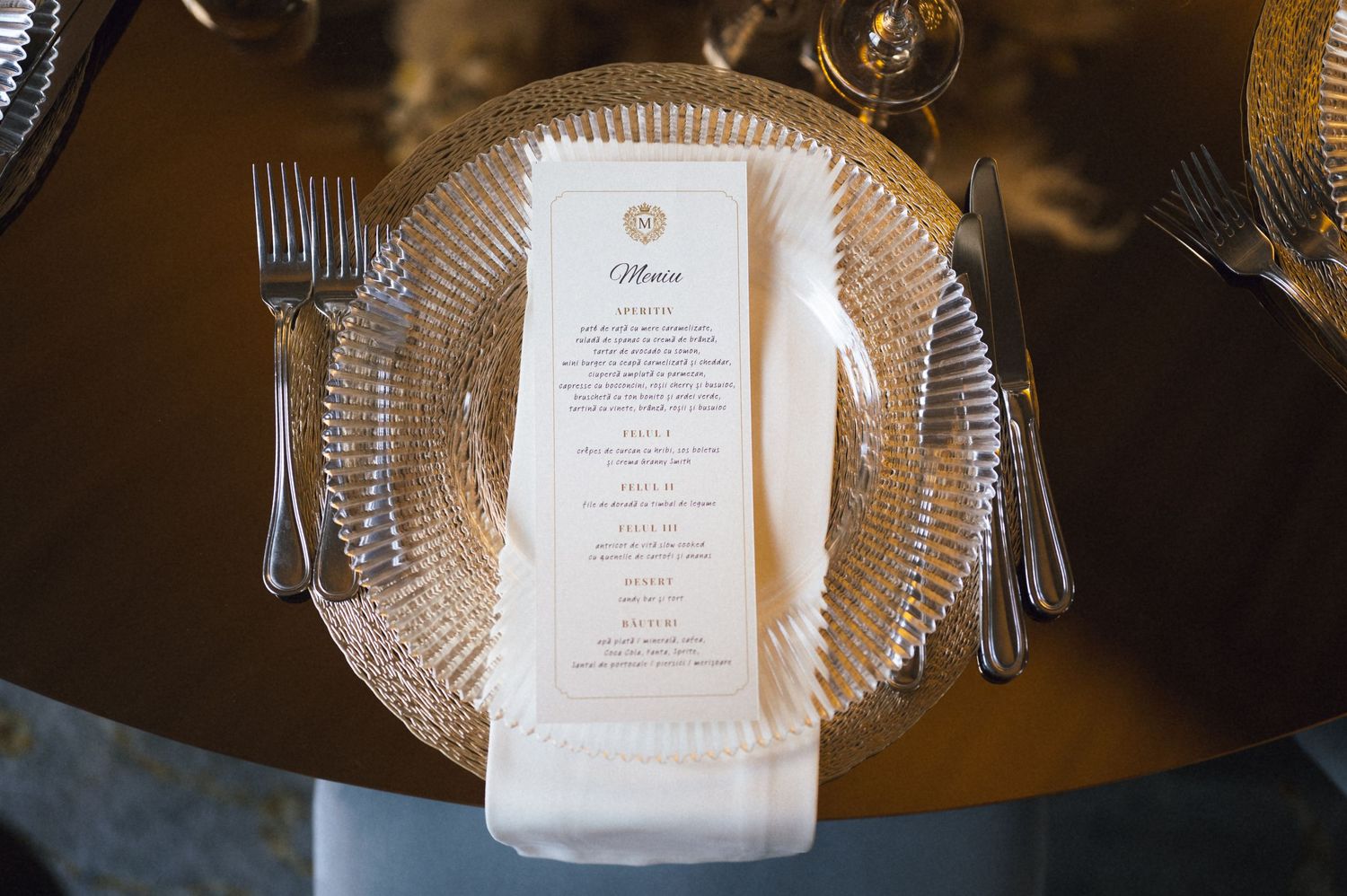 Elegant wedding place setting with crystal charger plate, white napkin, menu card, and silverware on a gold table.