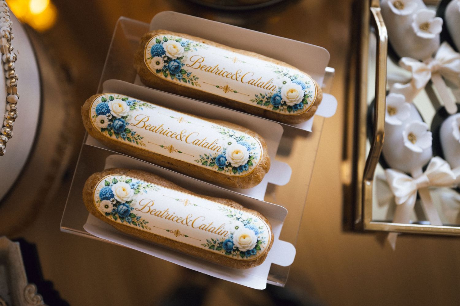 Elegant custom eclairs with blue floral designs and personalized names displayed in clear packaging at a wedding dessert table.