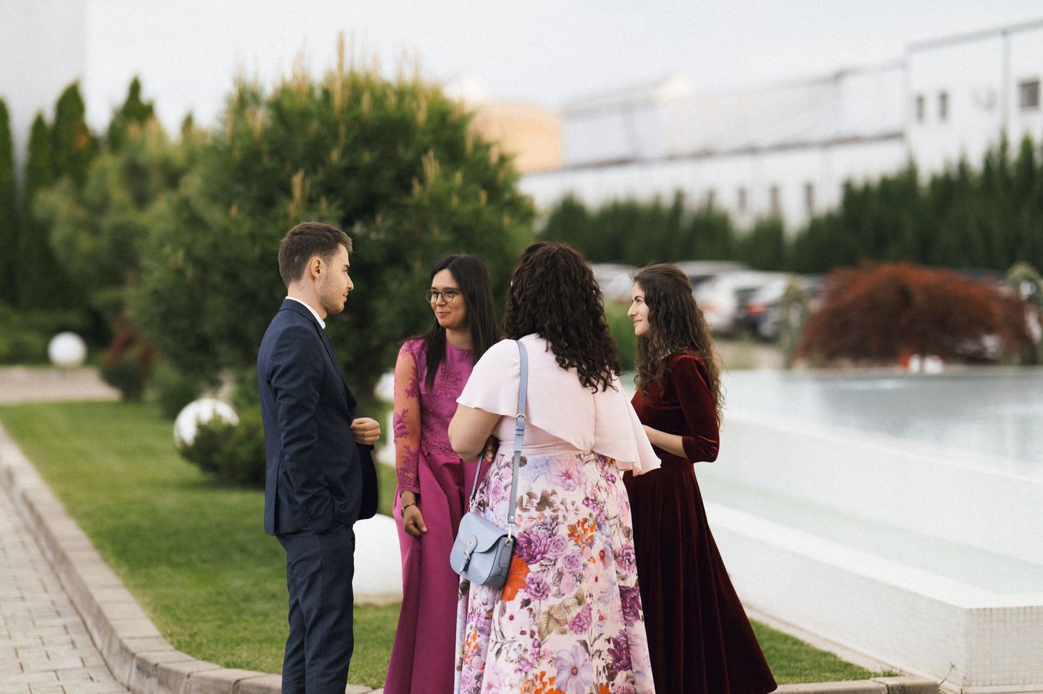 Elegantly dressed guests socializing outdoors at a formal event near a fountain and green landscaping.
