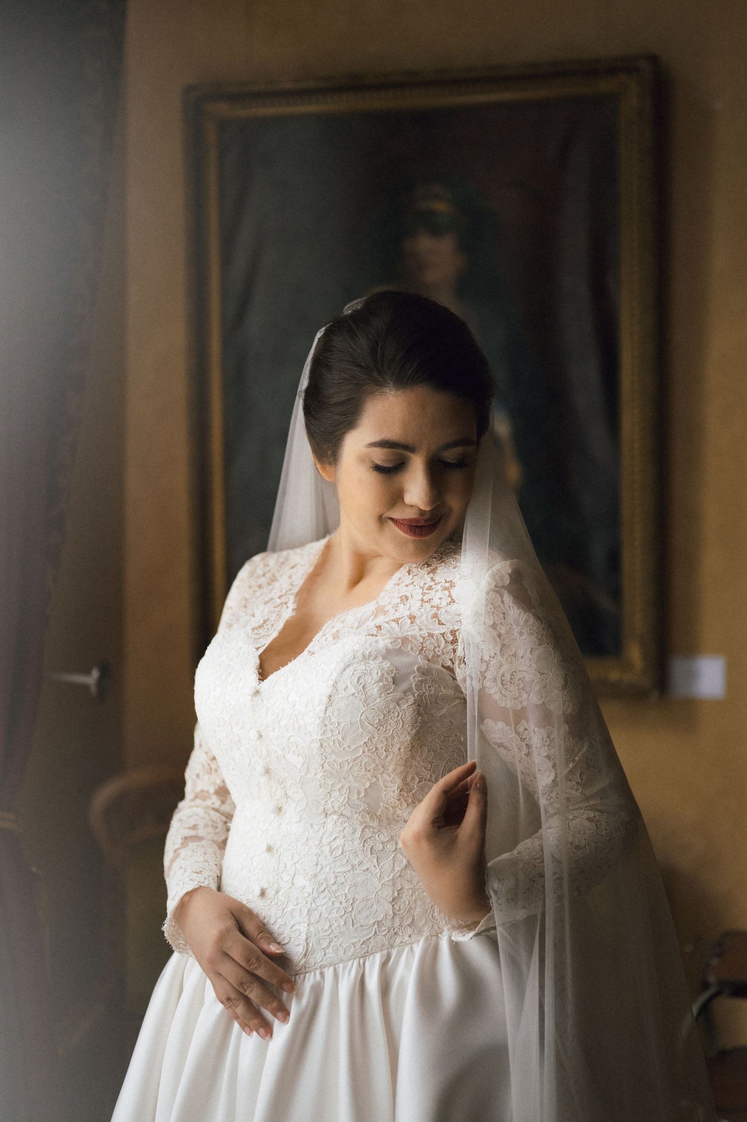 Bride in lace long-sleeve wedding gown and veil gazes down, posing elegantly before an ornate painting backdrop.