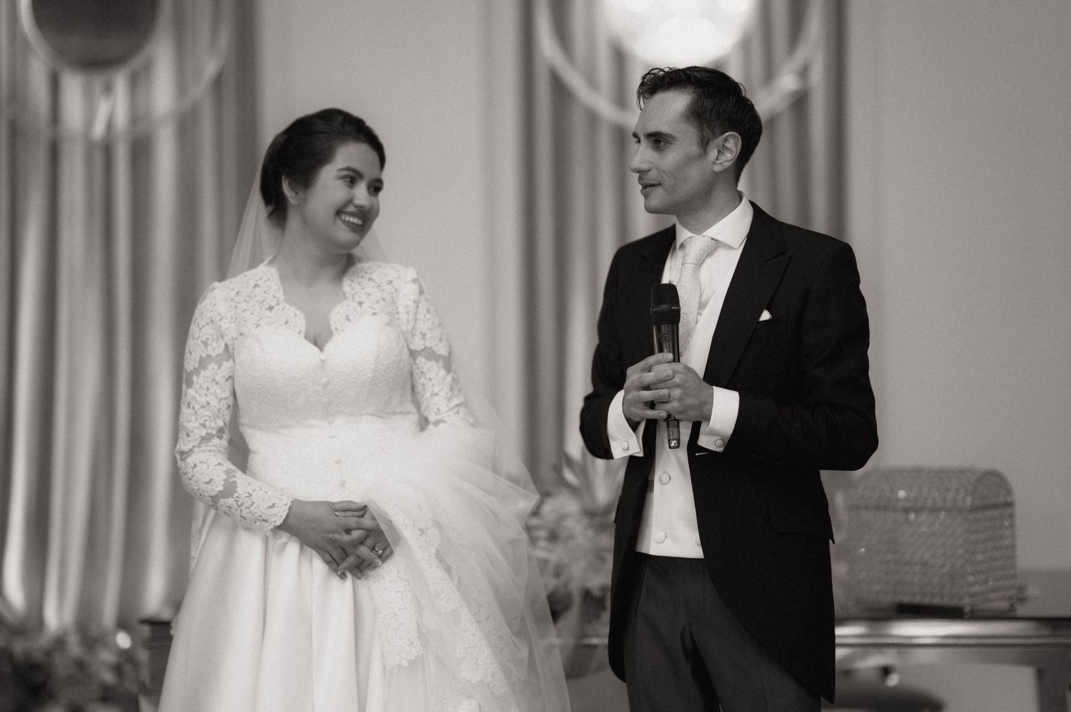 Black and white photo of a smiling bride in lace gown and groom in suit speaking at their wedding reception.