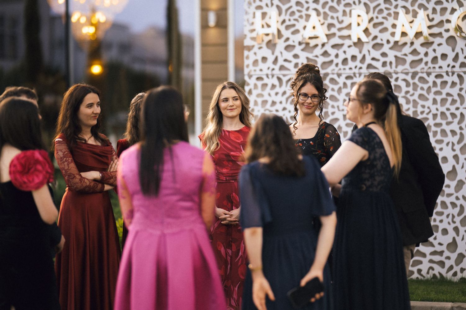 Elegantly dressed women in formal gowns socializing outdoors at an evening event near decorative signage.