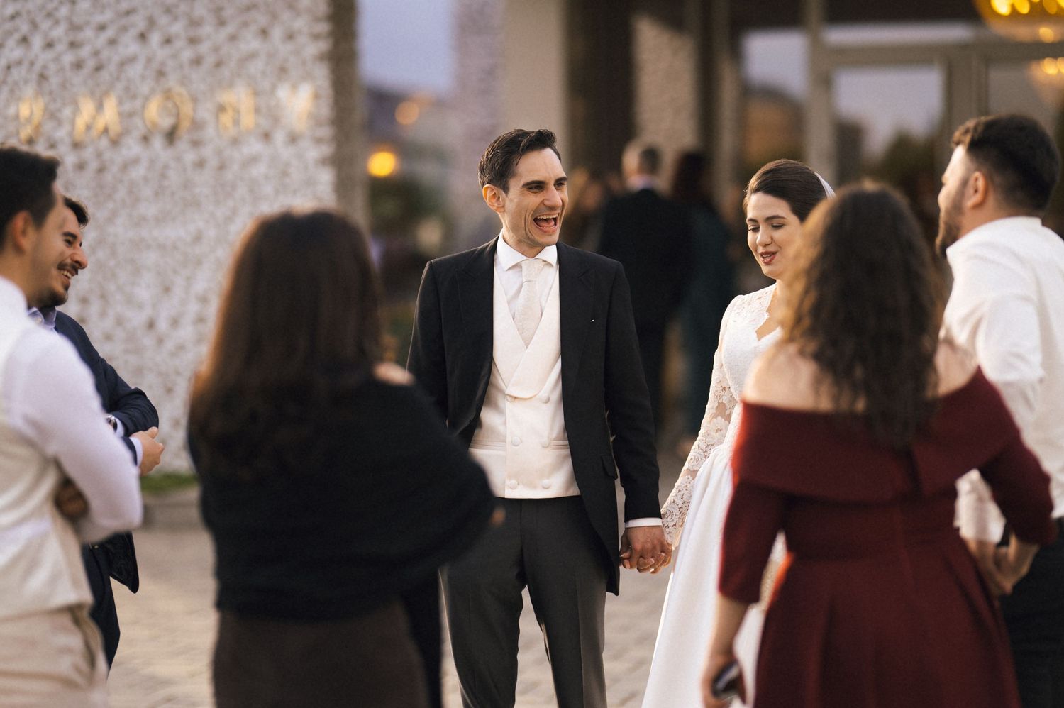 Bride and groom holding hands, smiling with wedding guests outside an elegant venue at dusk.