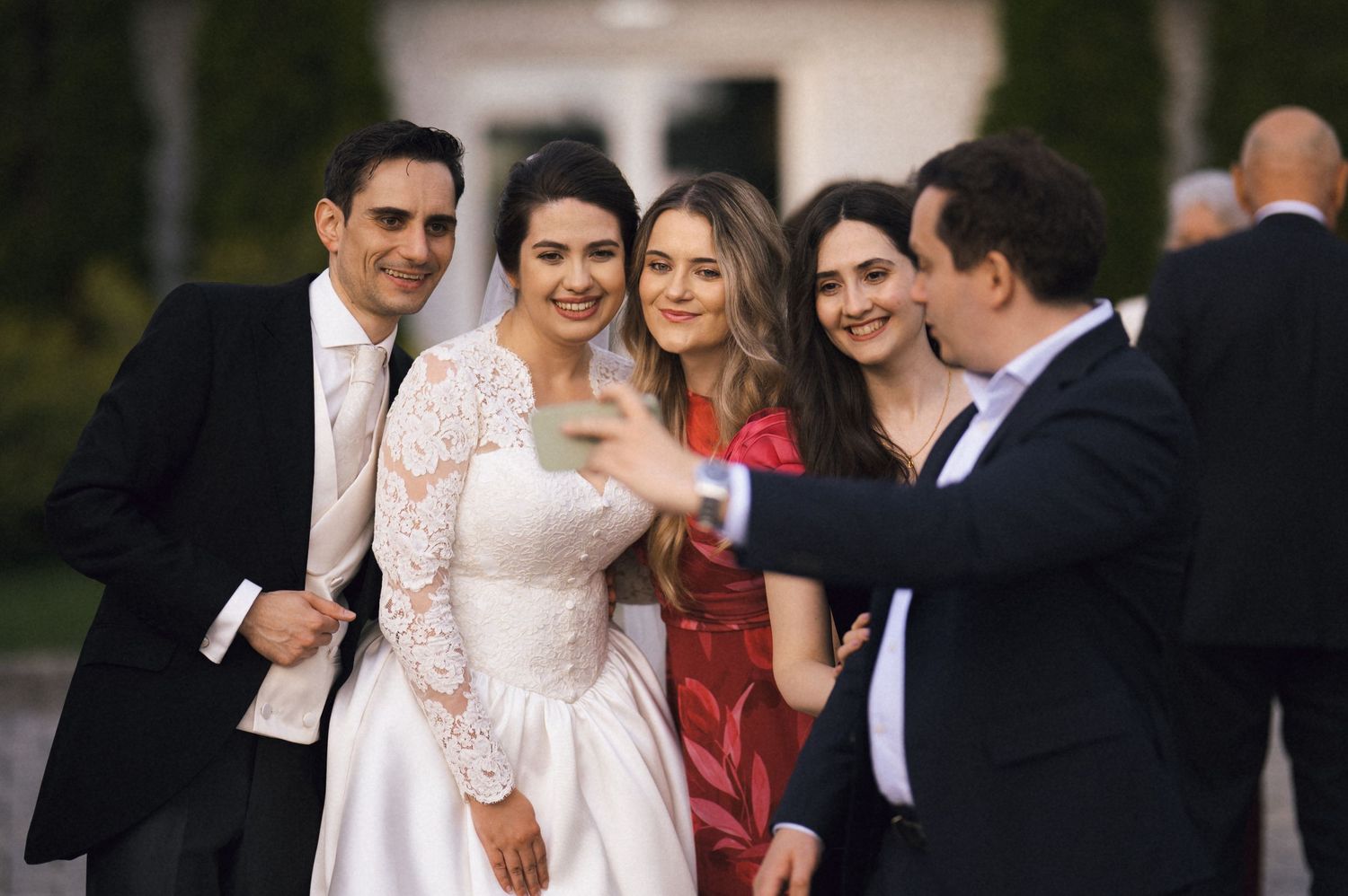 Bride in white lace gown poses with groom and guests for a selfie at an outdoor wedding celebration.