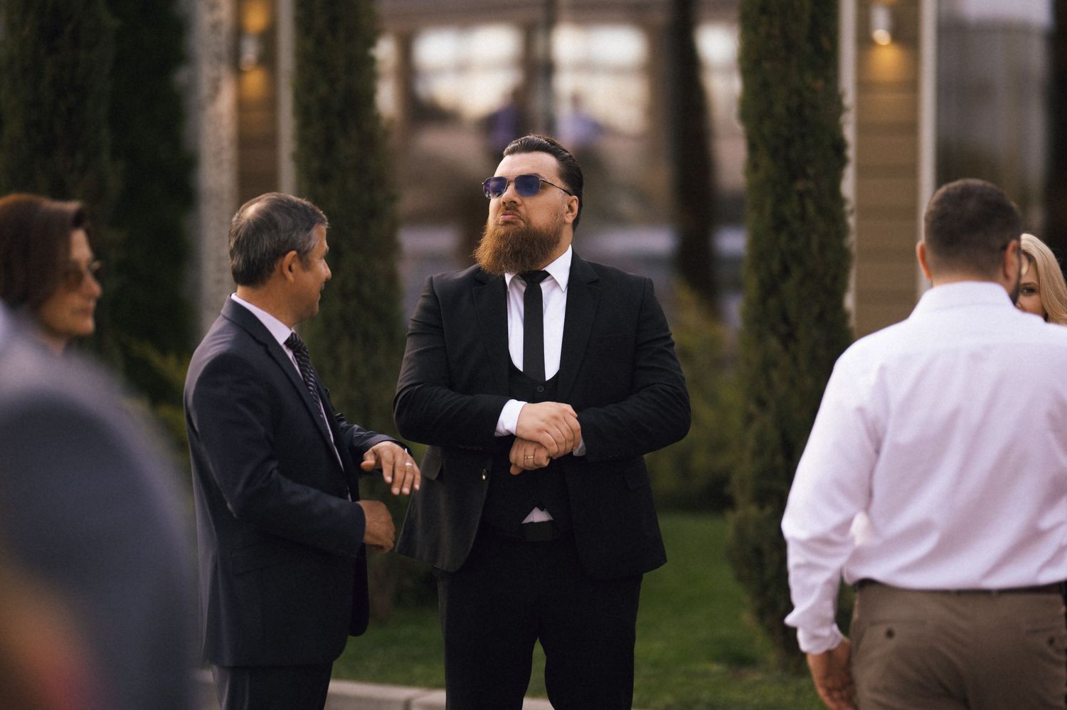 Men in black suits socializing outdoors at a formal event, with one wearing sunglasses.