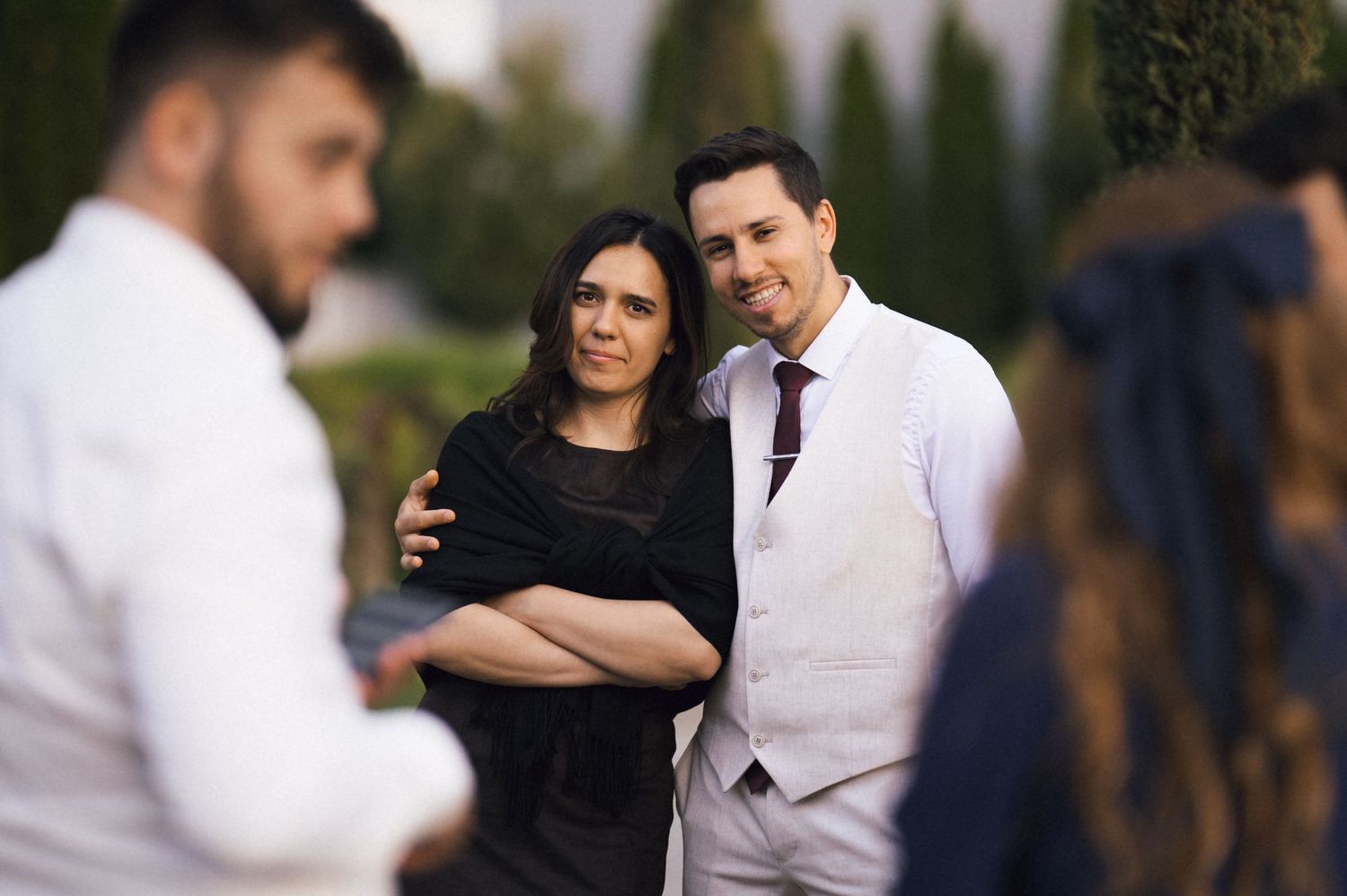 Couple posing outdoors at a wedding event, man in white suit with tie, woman in black outfit, smiling warmly.