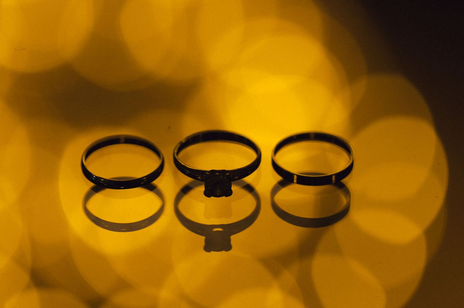 Three gold wedding rings with diamond engagement ring reflected on surface against warm bokeh golden background.