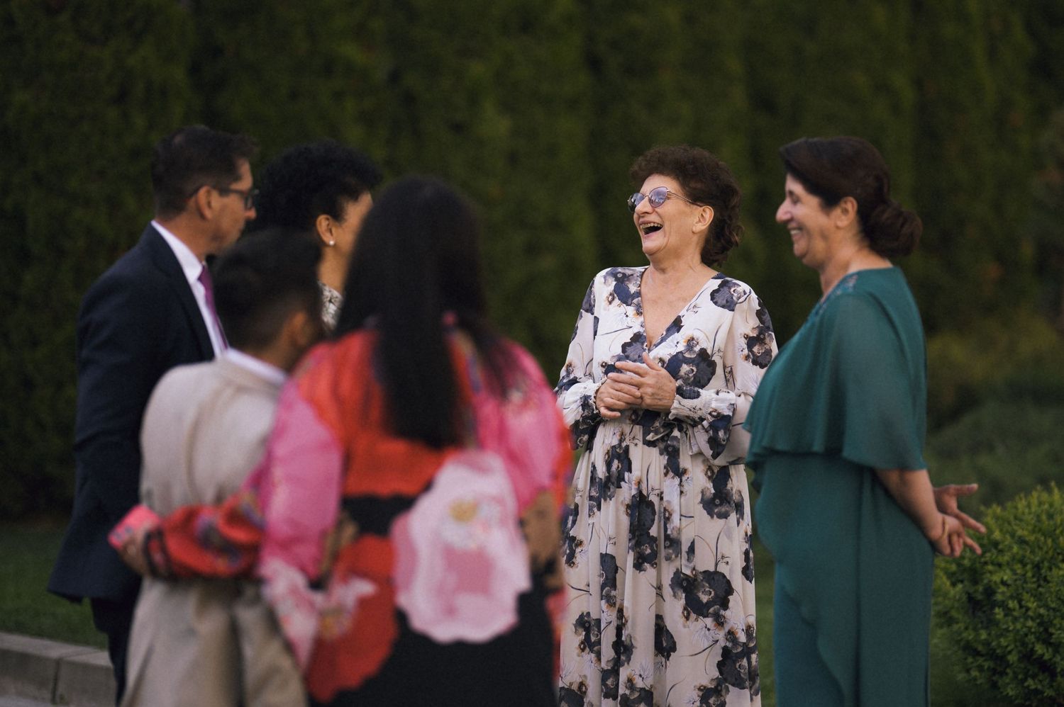 Guests laughing and socializing outdoors at a wedding reception, wearing elegant floral and green dresses.