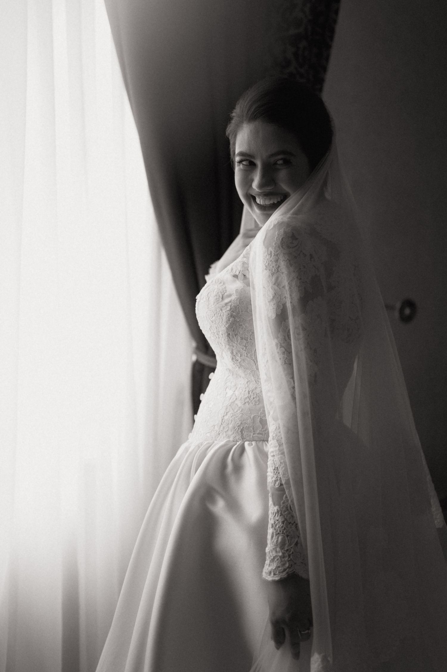 Elegant bride in long-sleeve lace wedding gown and veil smiles by window in black and white photo.