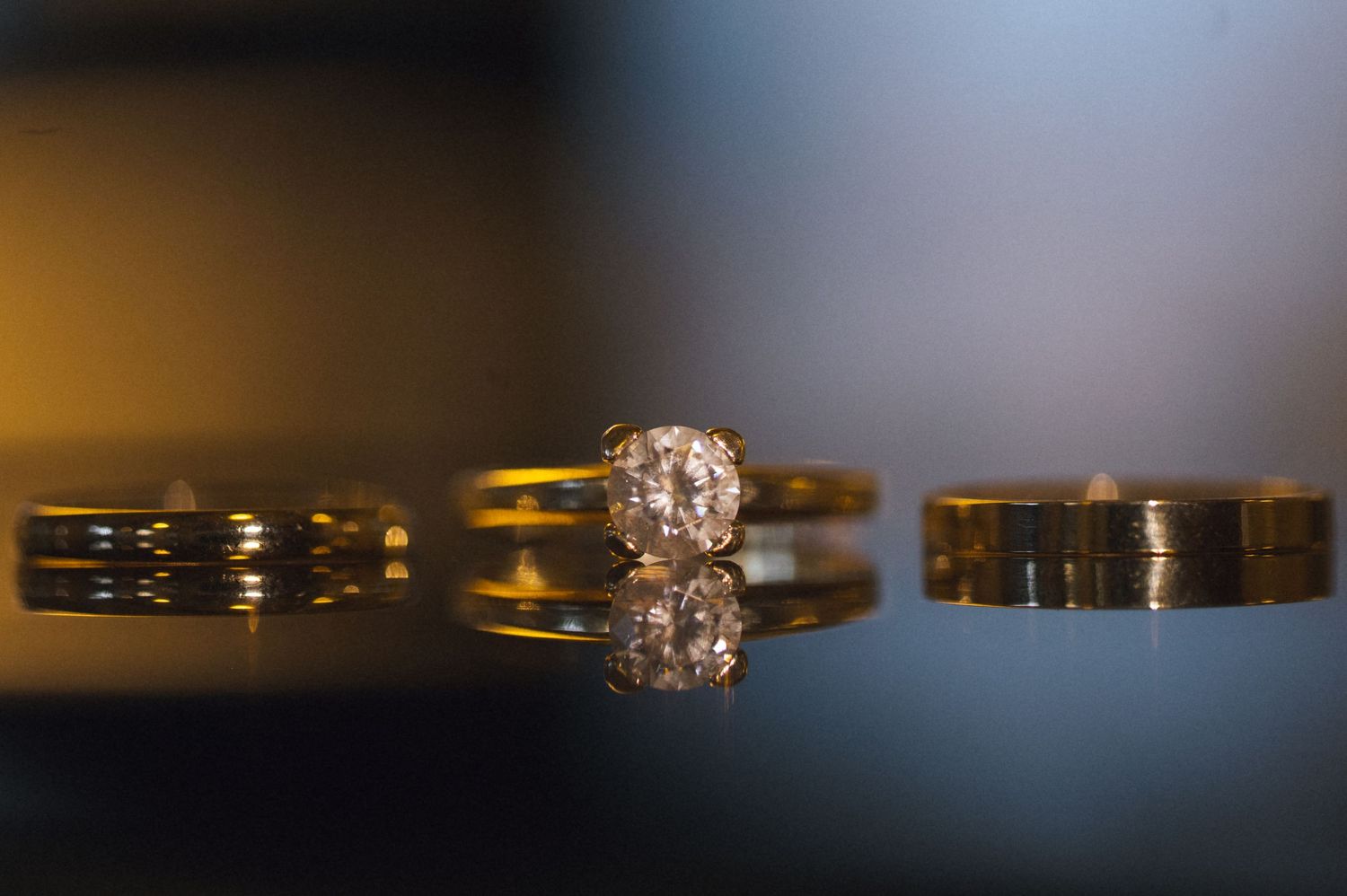Gold wedding bands and diamond engagement ring reflected on glossy surface with warm bokeh background.