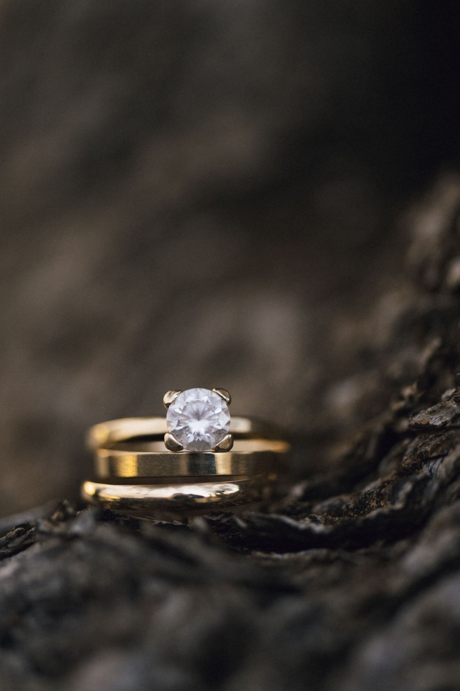 Gold wedding band and solitaire diamond engagement ring stacked on rustic dark wood surface.