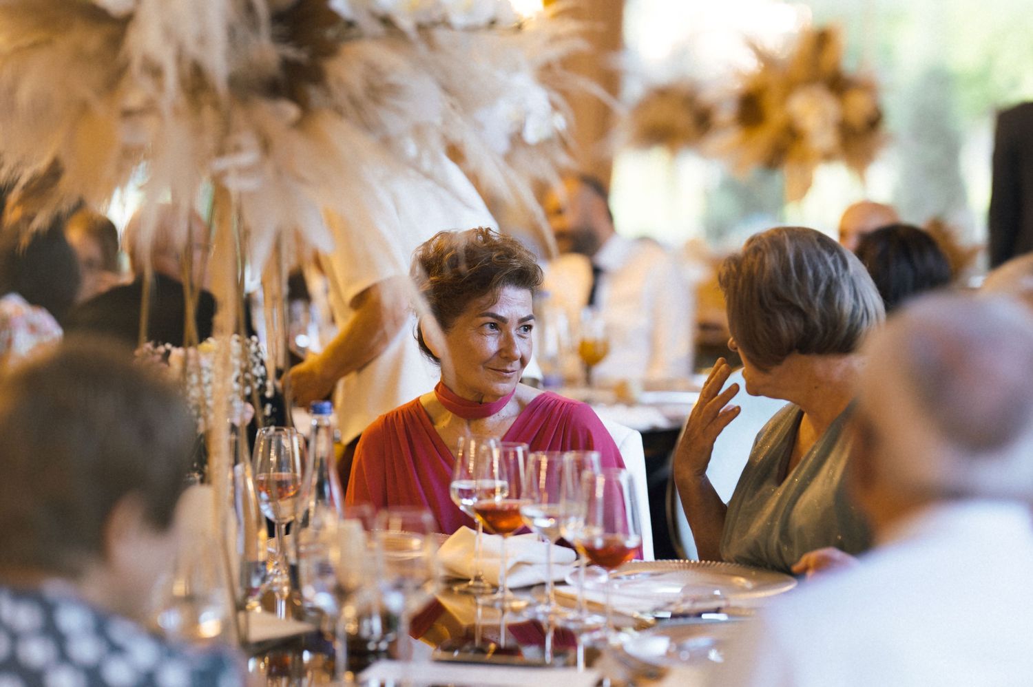 Guests enjoying lively conversation at elegant formal dinner with feather centerpieces and wine glasses on table.