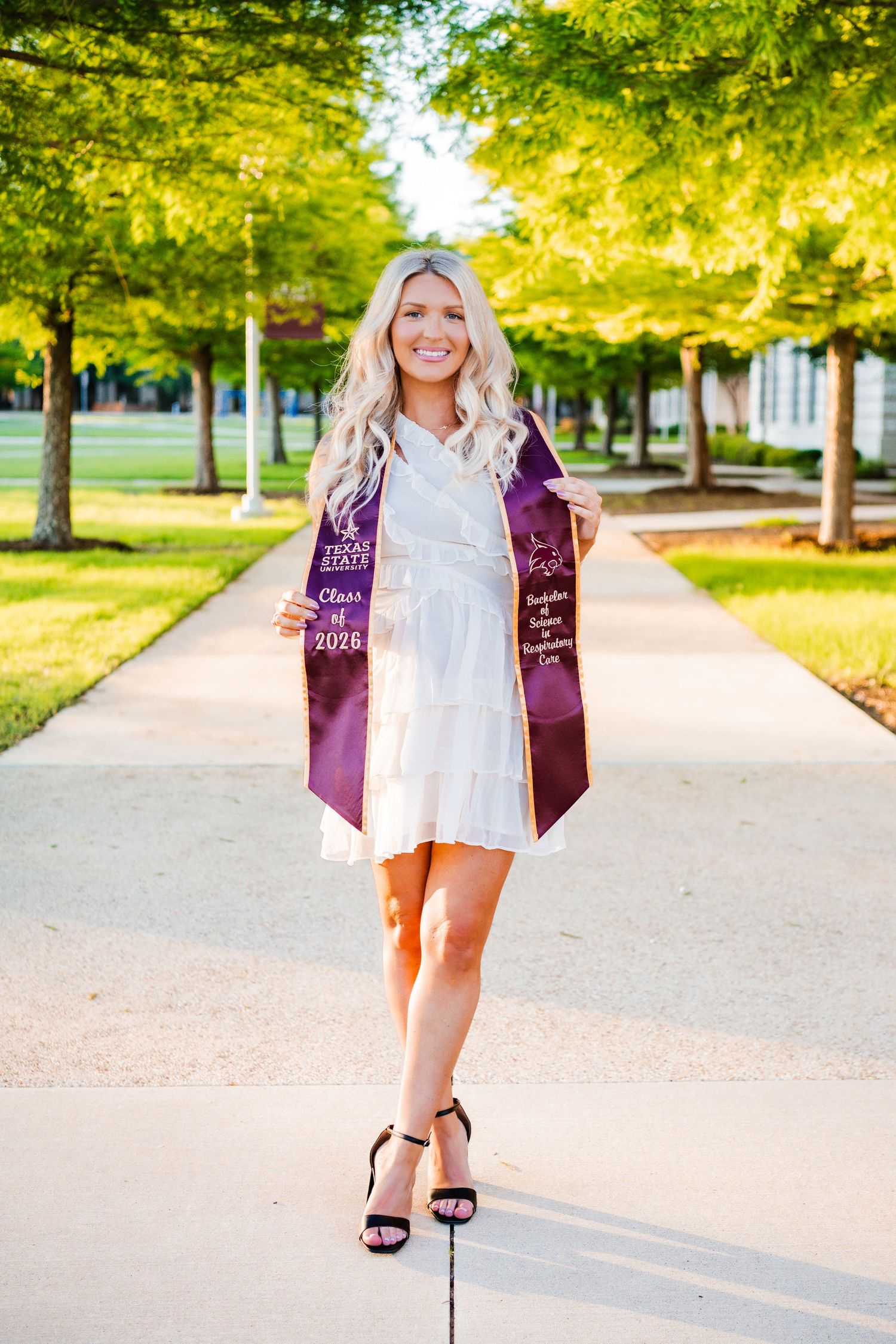Smiling blonde graduate in white dress holds purple Class of 2024 stole on a sunny campus walkway lined with green trees.