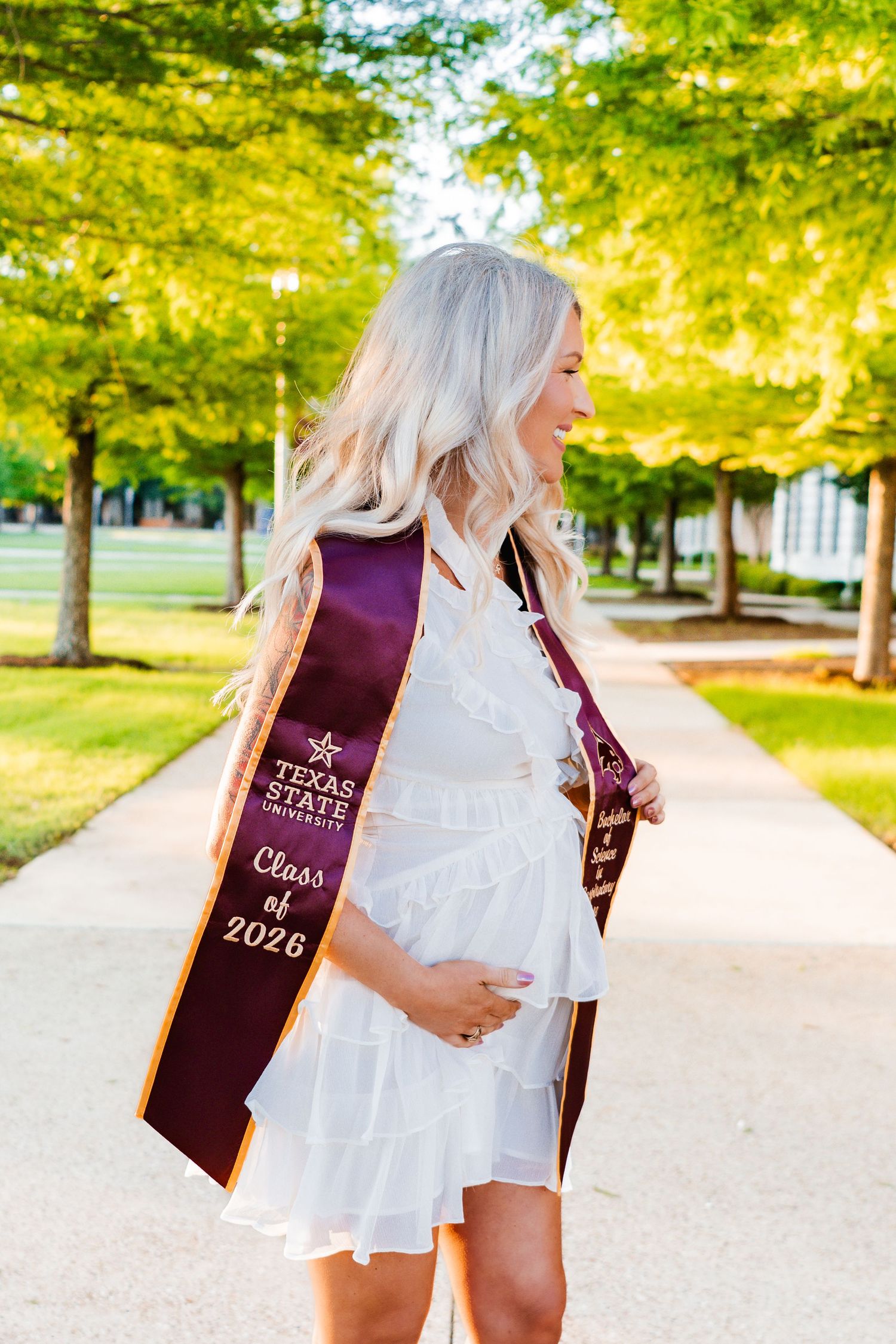 Pregnant woman wearing Texas State University Class of 2026 graduation sash in white dress on campus.