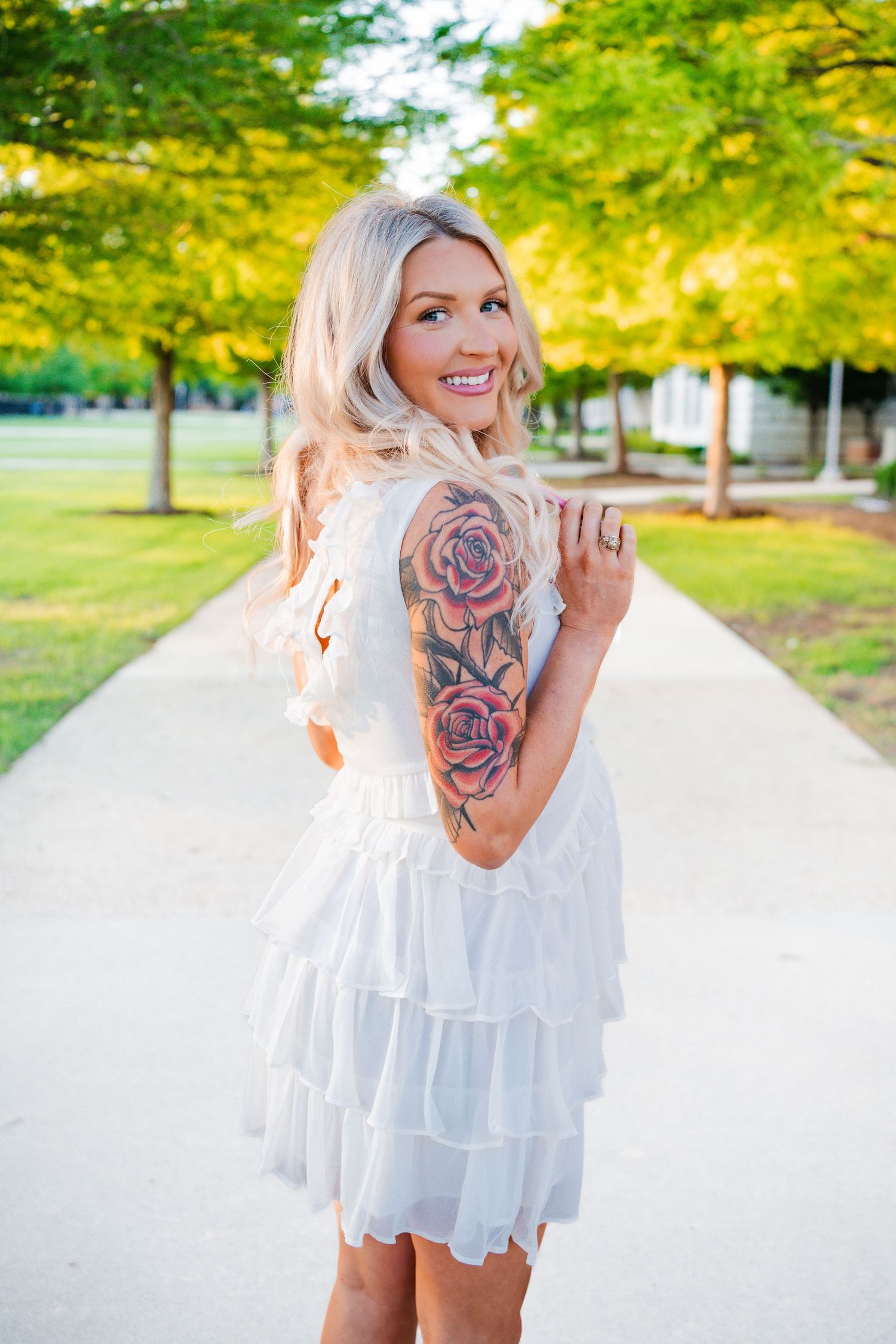 Smiling blonde graduate in white ruffled dress with rose tattoo sleeve poses on campus path.