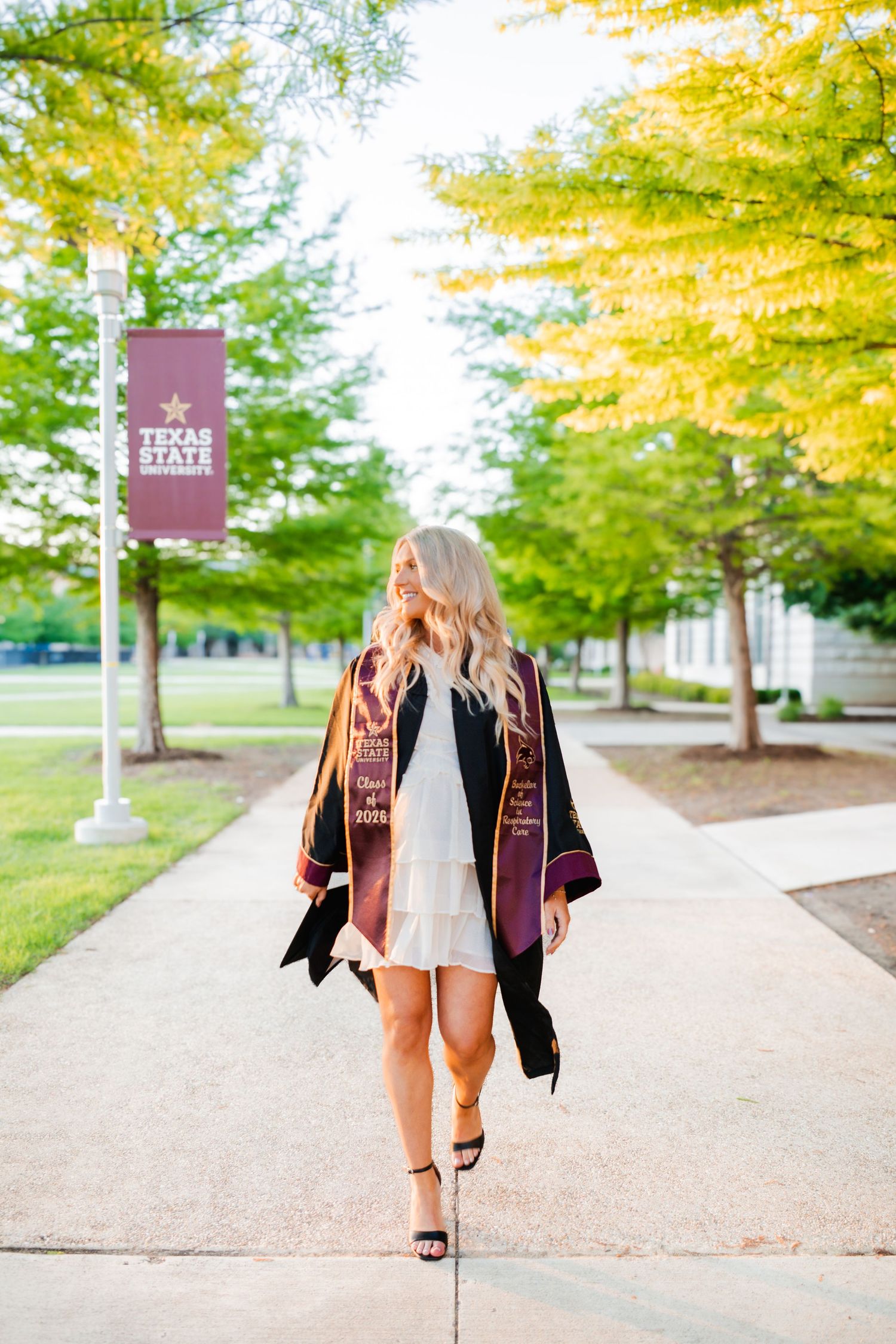 Blonde Texas State University graduate walking campus path in white dress, black gown, and maroon graduation stole.