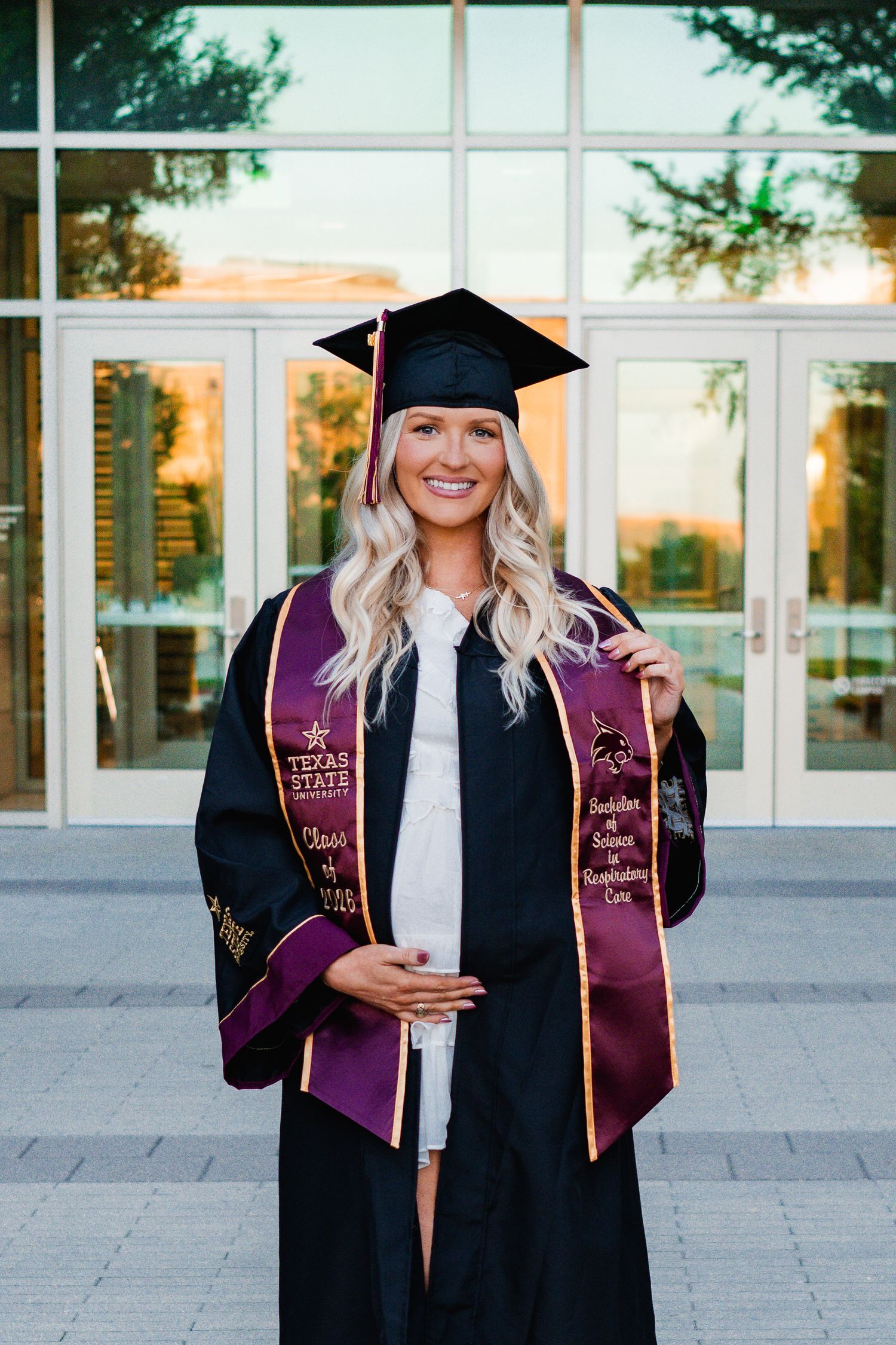 Smiling female graduate in black cap and gown with Texas State University maroon stole posing outdoors.