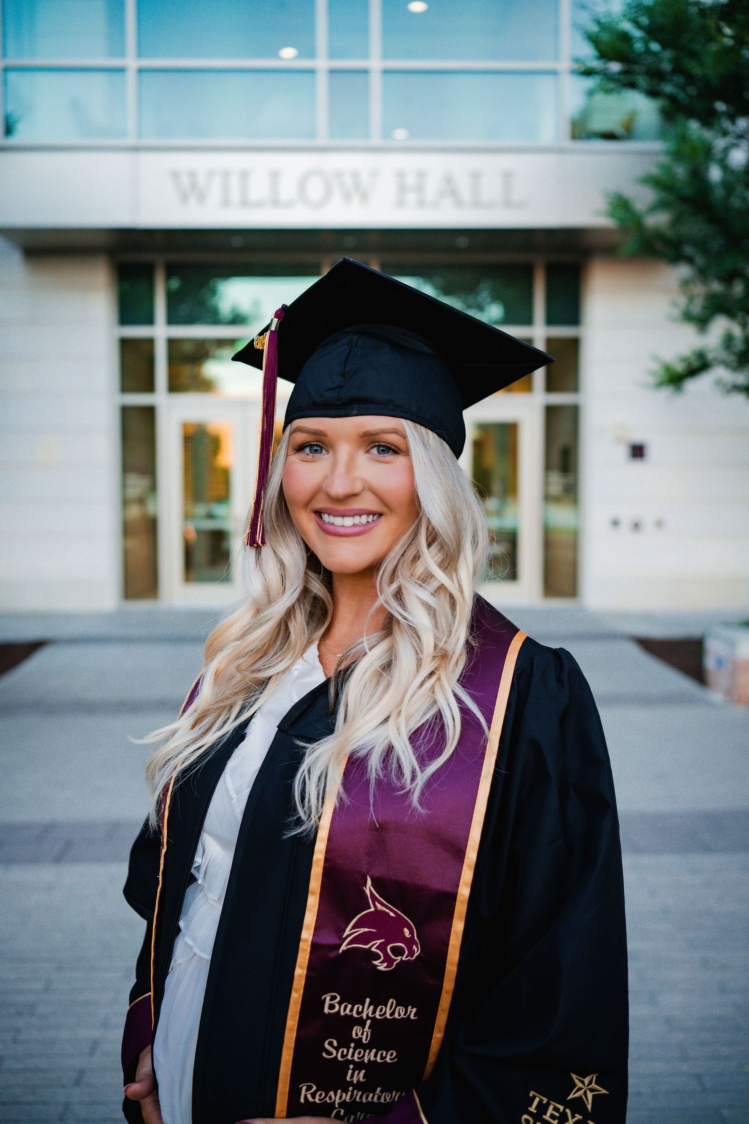 Smiling female graduate in black cap and gown with Bachelor of Science in Respiratory stole stands before Willow Hall.