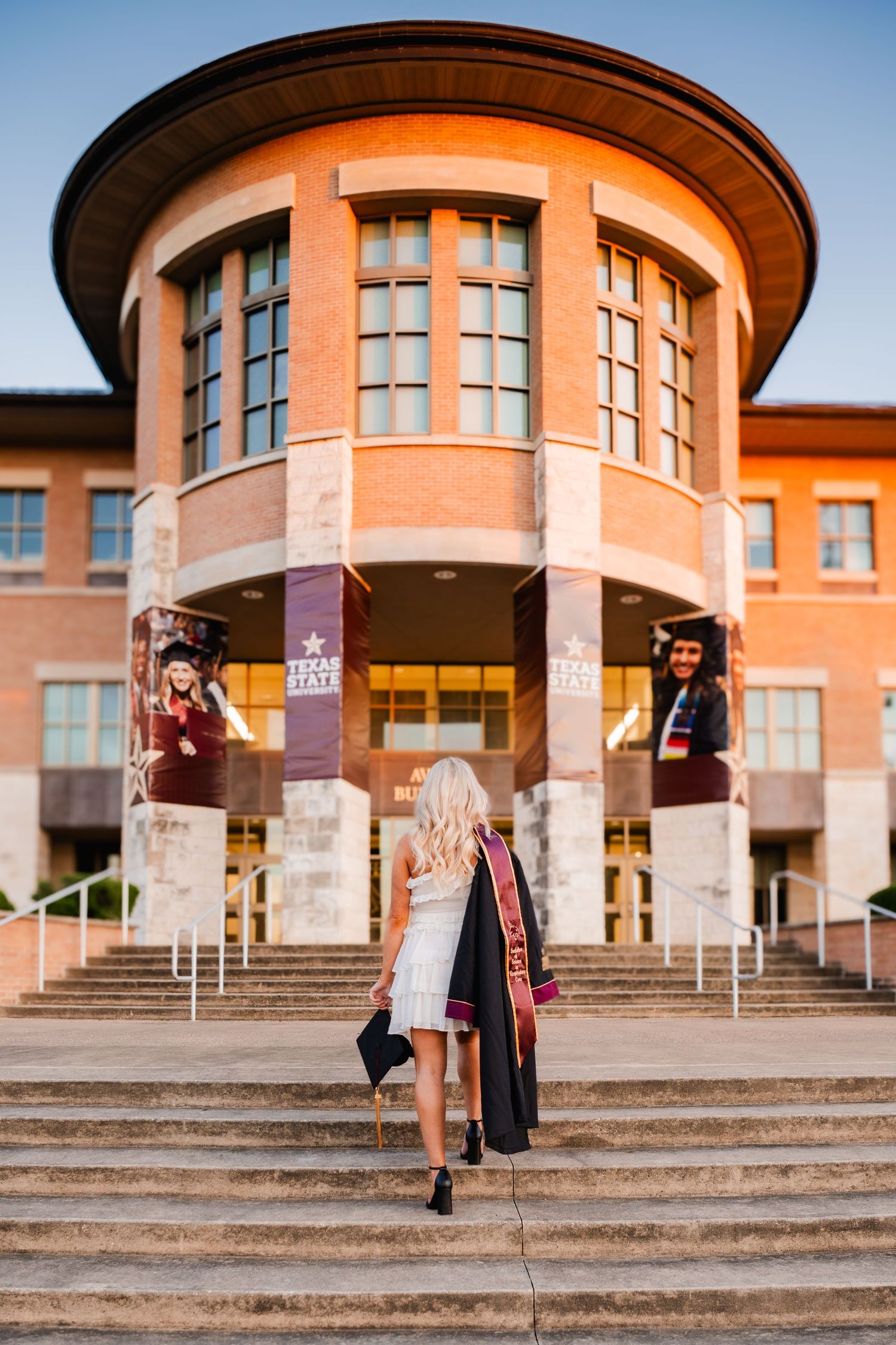 Blonde graduate in white dress and maroon sash walks toward Texas State University building with cap in hand.