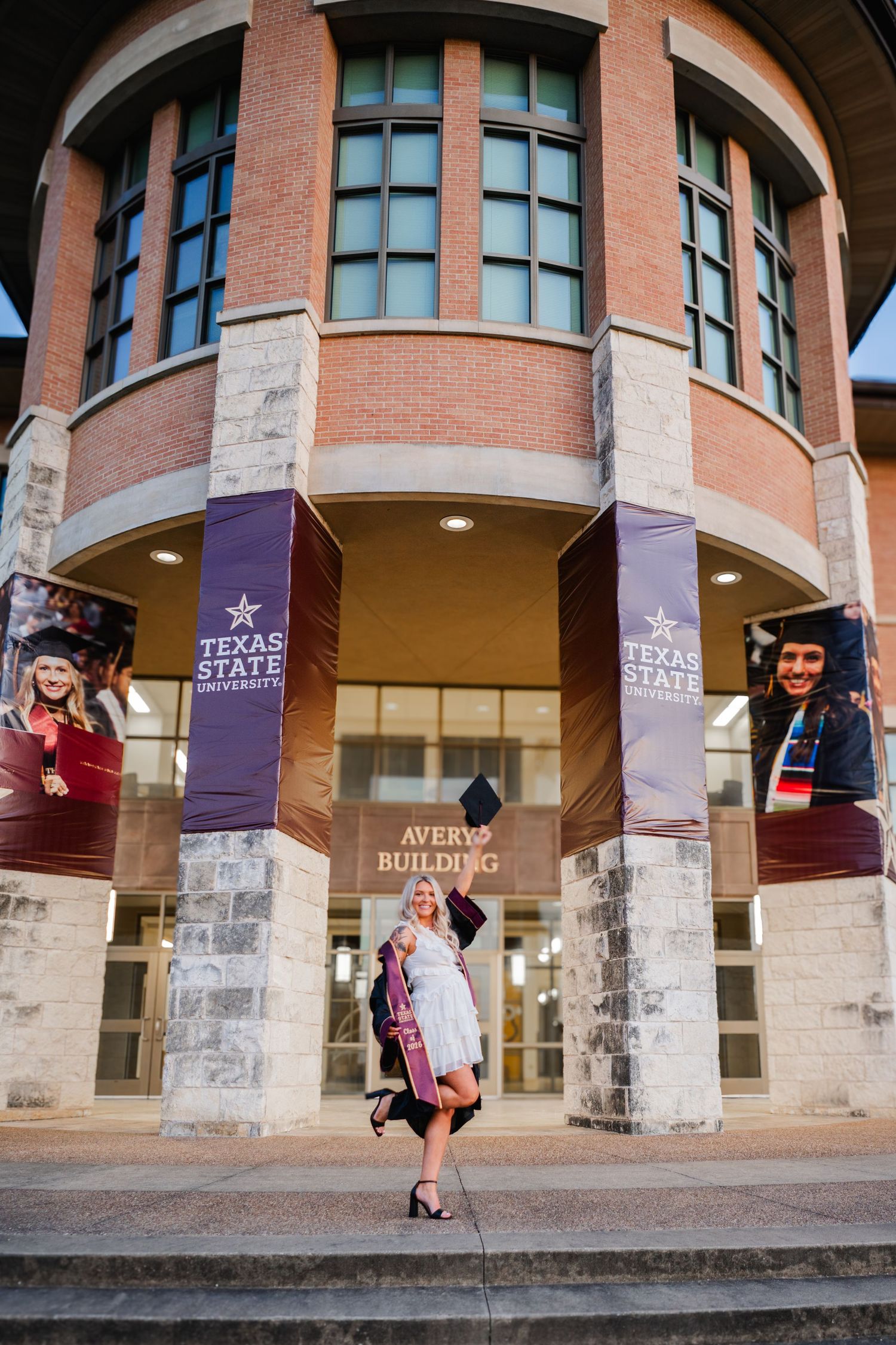 Graduate in cap and gown poses proudly in front of Texas State University Avery Building with purple banners.