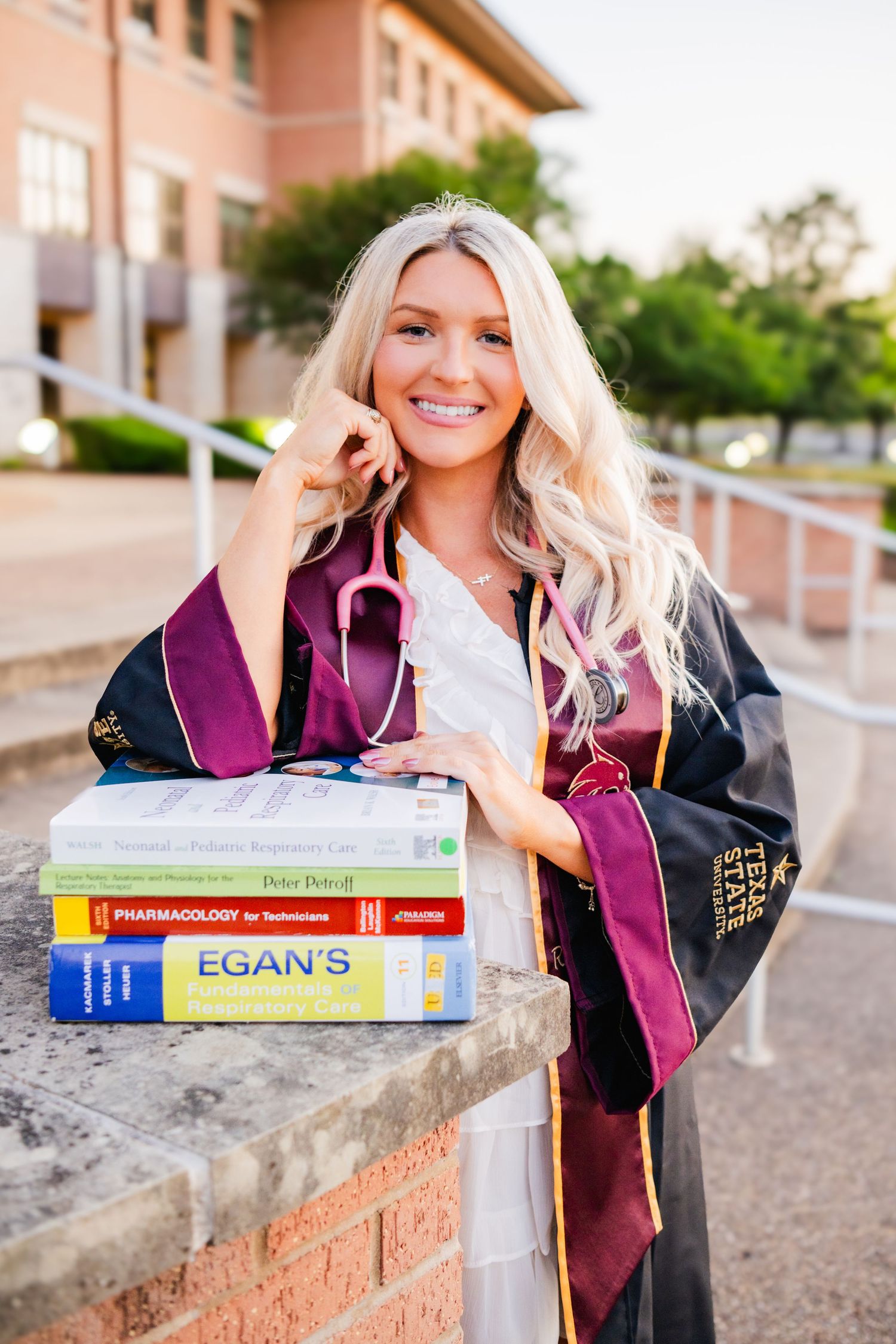 Blonde nursing graduate in black gown posing with medical textbooks including Pharmacology and Egan's on campus steps.