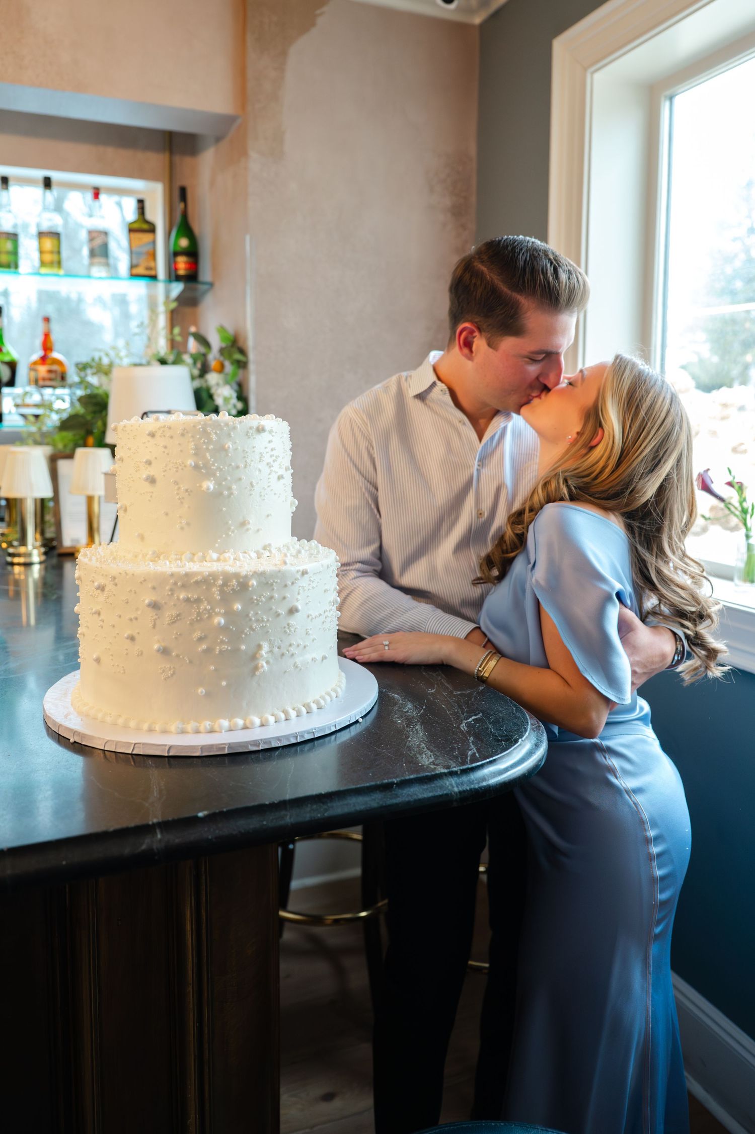 Couple kissing behind a white two-tier wedding cake on a dark bar counter in an elegant indoor venue.