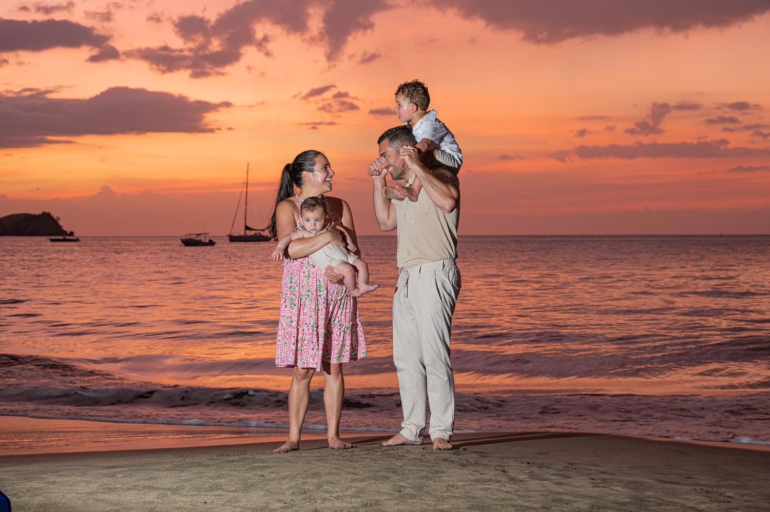 Family of four enjoying a beautiful orange sunset on a tropical beach with sailboats in the background.