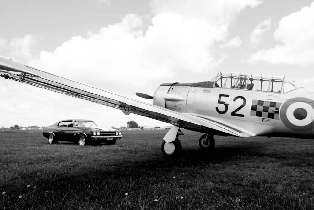 Black and white photo of a classic muscle car parked beside a vintage WWII trainer aircraft numbered 52 on a grassy airfield.