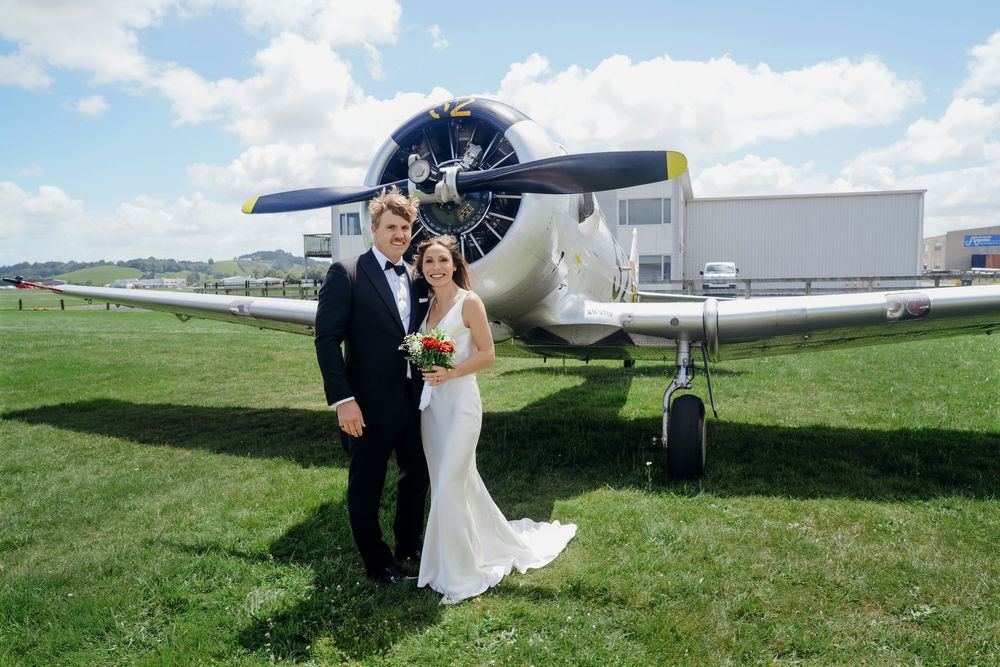 Bride and groom posing in front of a vintage propeller airplane on a green airfield on their wedding day.