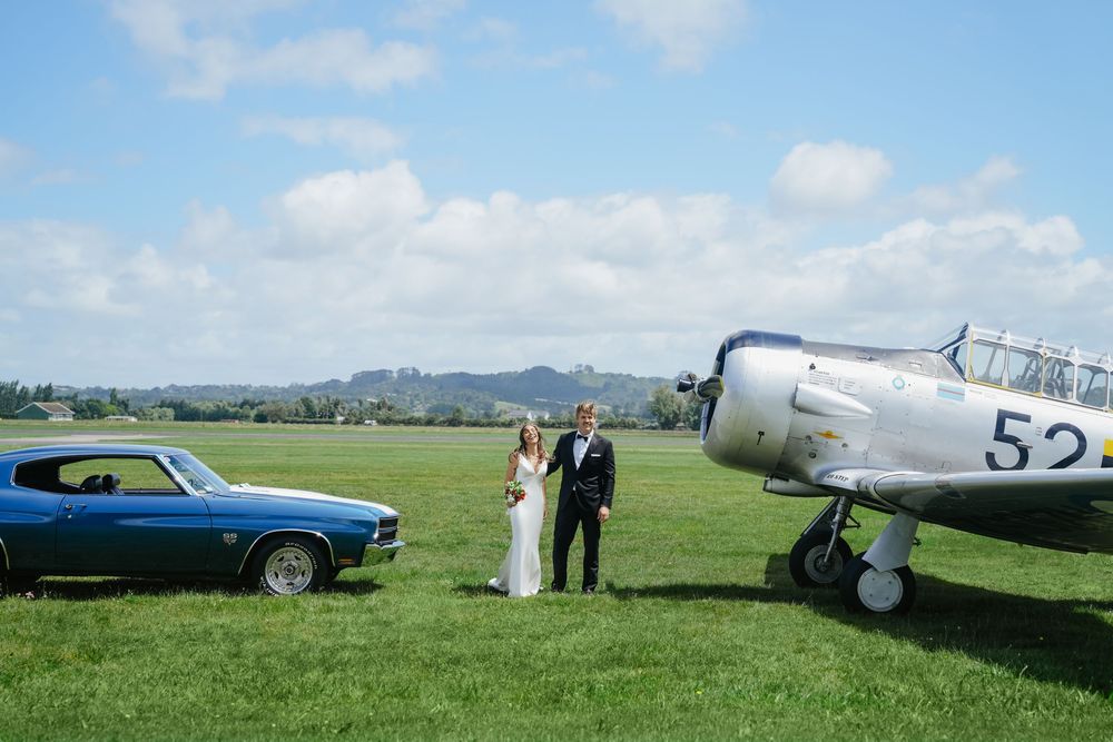 Wedding couple posing between a vintage blue Chevrolet Camaro and a classic silver Harvard aircraft on a green airfield.