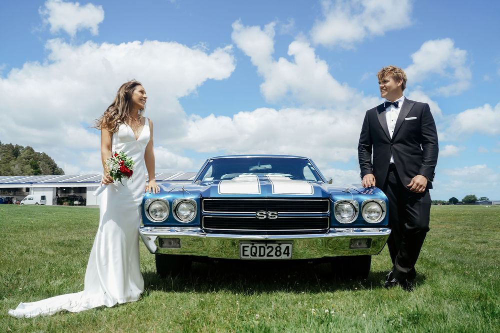 Bride and groom posing with a classic blue Chevrolet SS muscle car on a sunny day at their wedding.