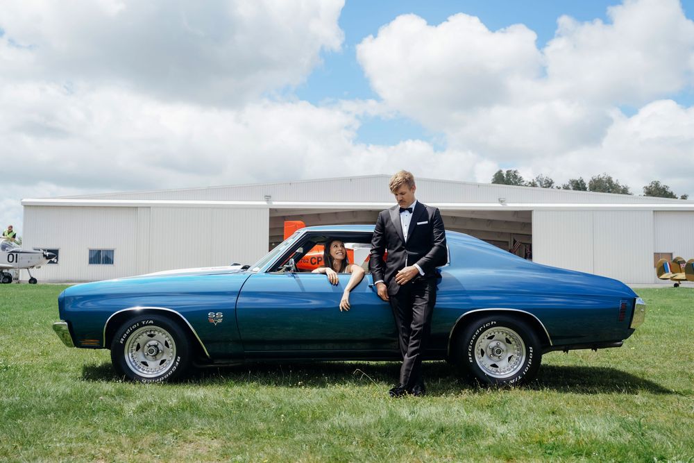 Groom in tuxedo leaning on a vintage blue Chevrolet SS while bride sits inside the classic car.