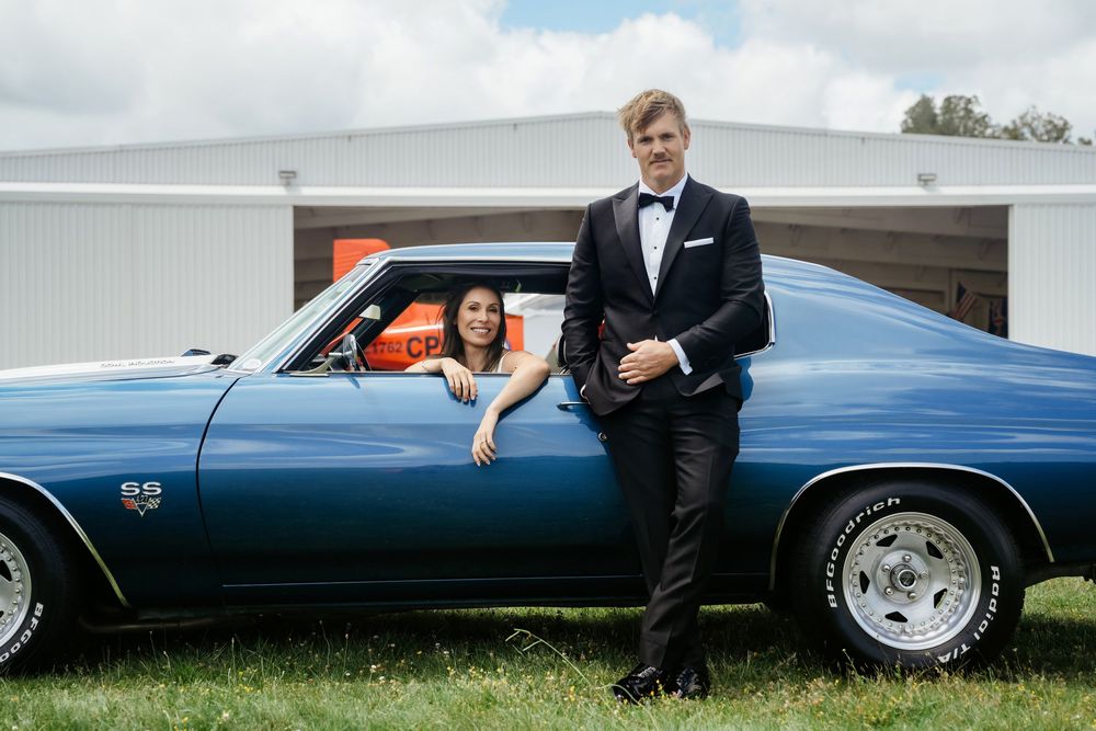 A couple poses with a classic blue Chevrolet SS muscle car outdoors, dressed in formal prom attire.