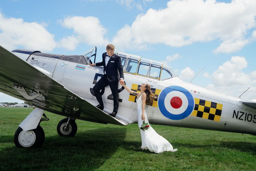 Bride and groom posing beside a vintage RNZAF warplane on a green airfield under a blue sky.