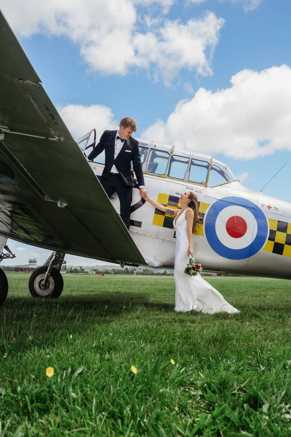 Bride and groom hold hands beside a vintage RAF roundel aircraft on a green airfield under a blue sky.