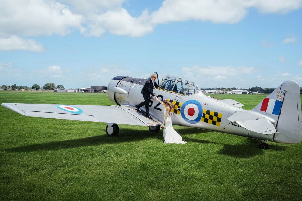 Bride and groom pose beside a vintage RAF Harvard aircraft on a green airfield under a blue sky.