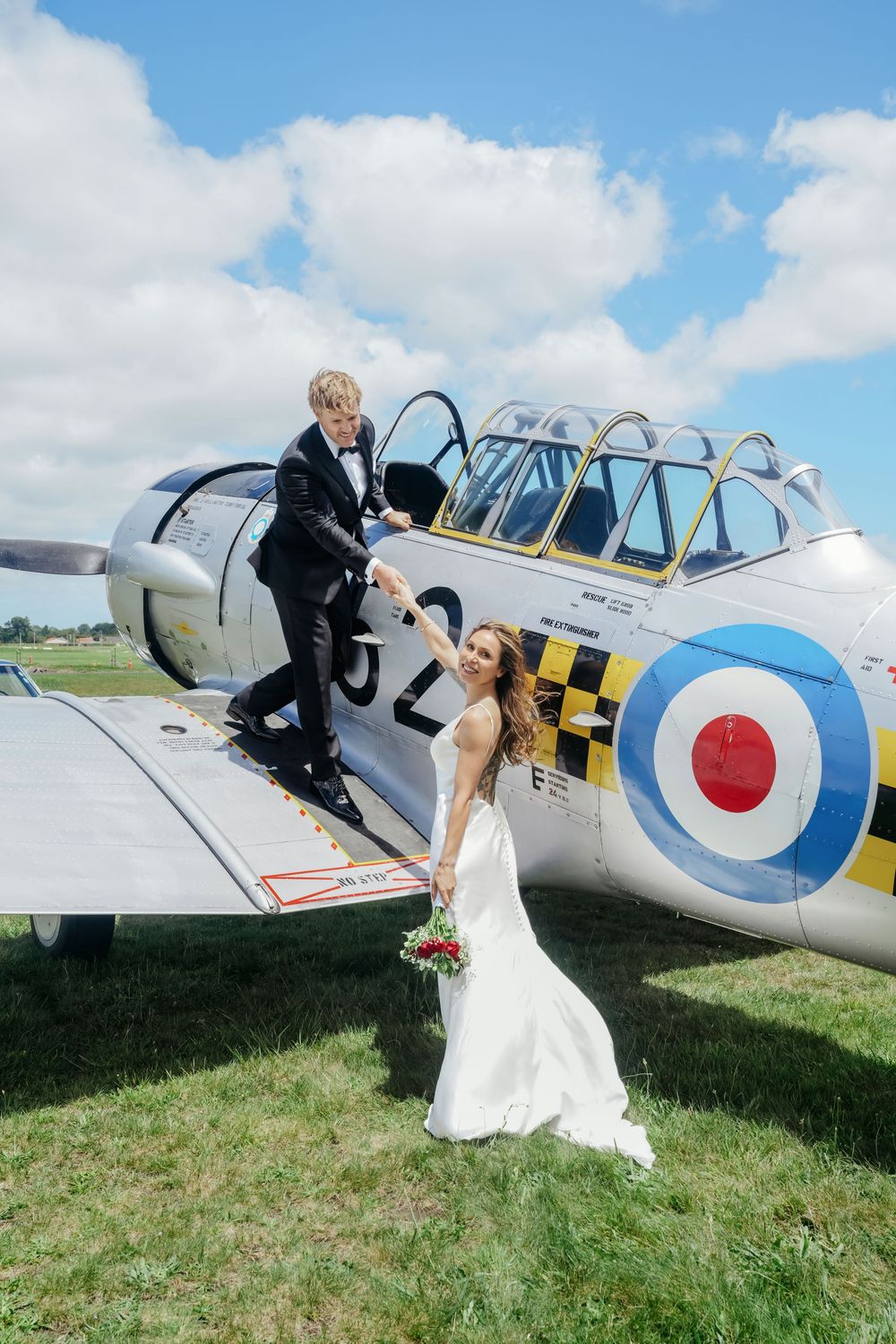 Bride and groom posing on a vintage RAF aircraft wing on their wedding day, with blue skies and green fields behind them.