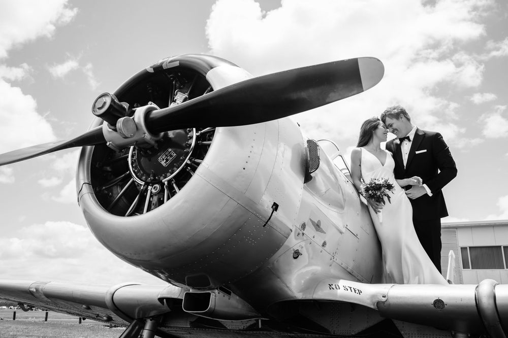 Black and white wedding photo of bride and groom kissing beside a vintage propeller airplane under cloudy skies.