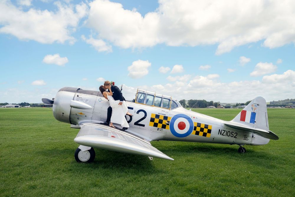 Couple kissing on a vintage WWII Harvard trainer aircraft NZ1052 on a green airfield under blue skies.