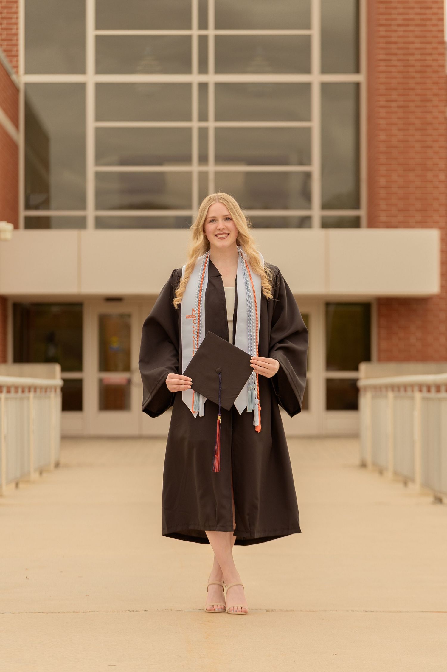 Smiling blonde graduate in black cap and gown holding diploma, posing in front of a brick university building.