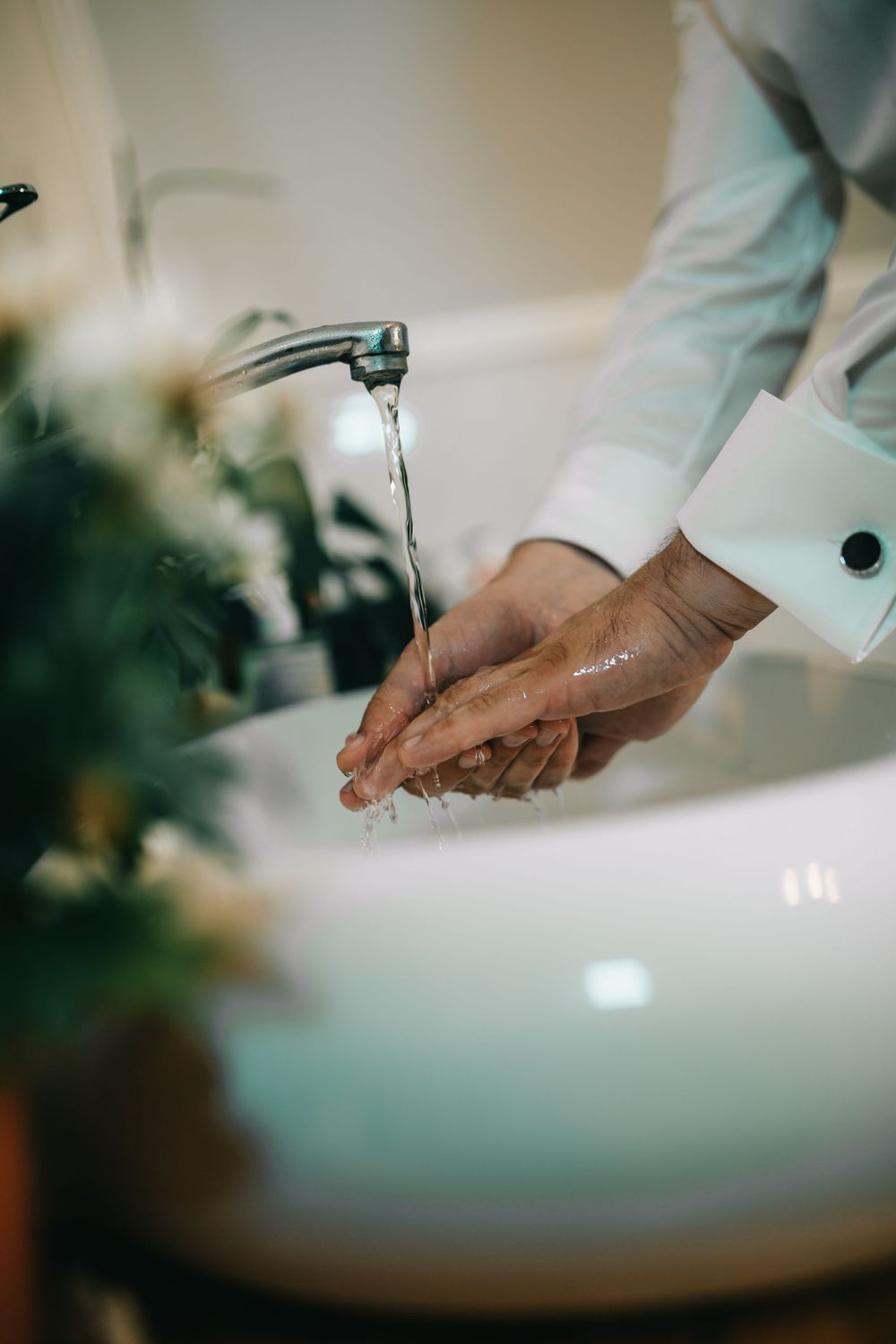 Person in white dress shirt with cufflinks washing hands under running faucet at white bathroom sink.